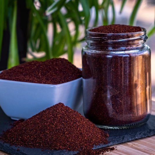 A clear glass jar filled with dark reddish-brown ground coffee sits next to a white bowl and a small pile of the same coffee granules on a dark surface, with blurred green leaves in the background.