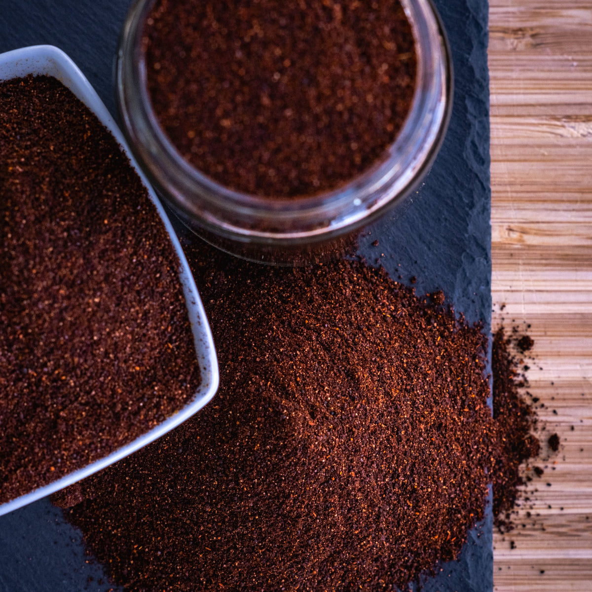 A close-up overhead shot of a dark brown ground spice or powder, shown in a clear glass jar, a white ceramic scoop, and spilled on a dark slate surface next to a bamboo cutting board.
