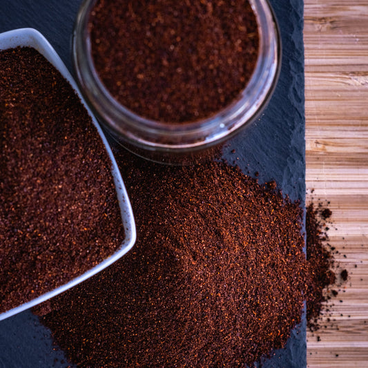 A close-up overhead shot of a dark brown ground spice or powder, shown in a clear glass jar, a white ceramic scoop, and spilled on a dark slate surface next to a bamboo cutting board.