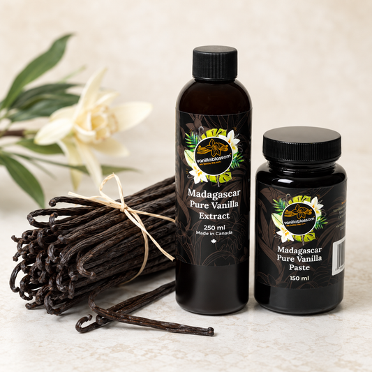 Two black bottles of vanilla extract and paste displayed with a bundle of vanilla beans and a white flower on a light beige surface. The larger bottle is labeled Madagascar Pure Vanilla Extract 250 ml, and the smaller jar is labeled Madagascar Pure Vanilla Paste 150 ml.
